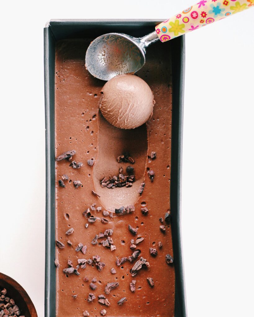 Delicious homemade chocolate ice cream in a tray with a colorful scoop, garnished with cacao nibs.
